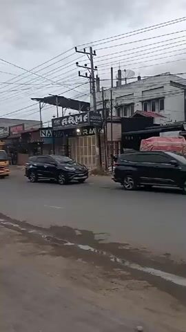 The video shows a street scene on an overcast day, likely in a town or city. Multiple vehicles are moving on an asphalt road that appears to have some dirt or mud on its edges.

In the foreground, a black sedan drives from right to left and out of frame. A black SUV travels in the opposite direction. Following these, a convoy of two yellow trucks with caged backs and a smaller yellow truck with a tarp-covered load drive from left to right. Another black SUV is seen on the right side of the road, moving away from the camera. A white truck is partially visible behind the yellow trucks.

The background features several buildings, suggesting a commercial area. Utility poles with numerous overhead power lines are prominent. Visible text on the buildings includes "EAT ARMA ARMAMI", "NADIA RM", "TREES", and "GUESS". The sky is entirely covered with clouds, indicating an overcast weather condition.