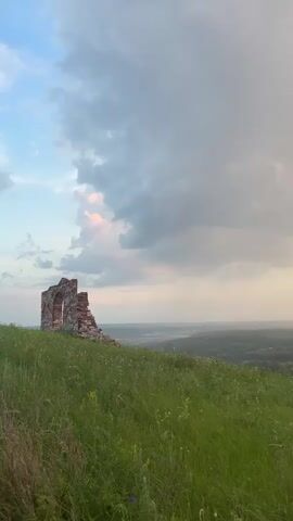 A video pans across a grassy hilltop with a ruined brick archway on the left. The horizon shows a lush green landscape with a winding river and a hazy sky with scattered clouds, some tinged with orange from the setting sun. There are no people or animals visible. The camera movement is slow and steady, capturing the expansive view.