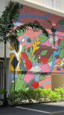 A colorful mural is painted on the side of a building. The mural features abstract shapes and depictions of plants and fruits in bright colors such as pink, green, red, and yellow. A palm tree stands to the left of the mural, with its fronds partially obscuring the artwork. In front of the mural, there is a line of green bushes. A paved road is visible in the foreground, and a light-colored car is parked partially in view on the right side of the frame. The sky is not visible. The time code indicates that the video is approximately 5 seconds long.