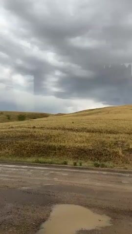 The video shows a view from a vehicle traveling on a dirt road. The road is wet and has several puddles. The landscape is a rolling hill covered in dry grass. The sky is overcast with dark, heavy clouds.
