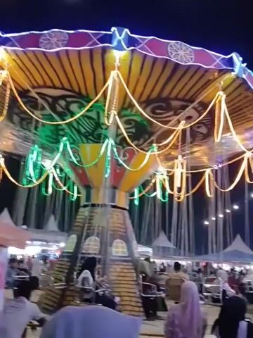 The video captures a lively night scene at what appears to be a fairground or carnival. In the foreground, a swing ride with colorful lights and decorative elements is prominently featured. People are visible sitting on the swings, suggesting the ride is in motion or preparing to operate. In the background, a large Ferris wheel with intricate, illuminated patterns glows against the dark night sky. Numerous tents and stalls can be seen, indicating a bustling marketplace or event area. A crowd of people, many wearing hijabs and traditional attire, are milling about, enjoying the festivities. String lights are hung throughout the area, further illuminating the scene and adding to the festive atmosphere. The overall impression is one of vibrant activity and community gathering under the night sky.