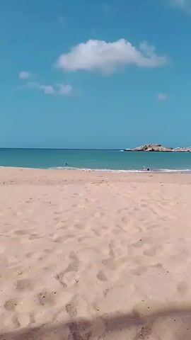 The video shows a wide shot of a beach with light-colored sand under a partly cloudy blue sky.  Several people are visible on the beach and in the water.  There are some rocks or a small island in the distance, situated offshore.  As the camera pans slightly right, it reveals a lifeguard tower and some trees.  The shadows of trees are cast onto the sand in the foreground.