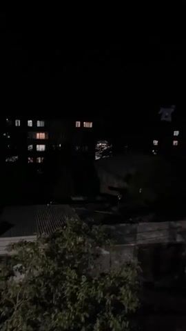 The video is a nighttime shot of a dimly lit urban environment. Buildings with illuminated windows are visible in the background. In the foreground, there are trees and some structures with corrugated metal roofs. The lighting is dark, with artificial light sources from the buildings casting a warm glow. The camera appears to be panning slightly or is handheld, causing minor movements. There are no people or animals visible. The overall atmosphere is quiet and nocturnal.