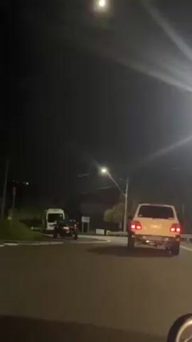 At night, a white SUV performs a burnout on a curved road, emitting smoke behind it. In the background, a white van is parked, and a dark car is parked closer to the viewer. The scene is lit by streetlights, with a bright moon visible in the sky. The SUV's brake lights are illuminated.