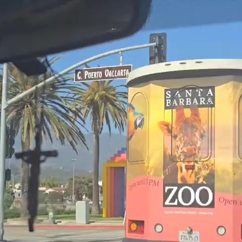 A car drives down a street in Santa Barbara, California. A sign overhead says "C. Puerto Vallarta." There are palm trees on the side of the road. A colorful archway made of concentric rectangular blocks painted in the colors of the rainbow sits in the distance, partially obscured by a bus advertising the Santa Barbara Zoo. The bus shows pictures of animals and the zoo's hours. A crucifix hangs from the car's rearview mirror. There is also the sound of music playing.