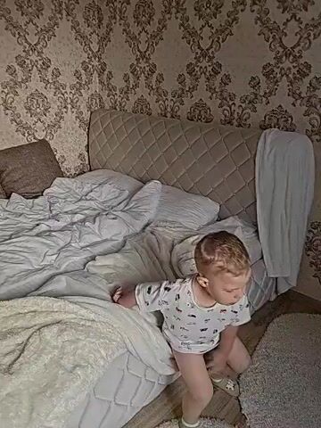 A child in pajamas is on the floor in a bedroom. The child is sitting on a white rug. There is a white potty next to the child. The child is looking at the camera. There is a bed behind the child. There is a patterned wallpaper on the wall behind the bed. There is a nightstand with a lamp on it to the right of the bed.