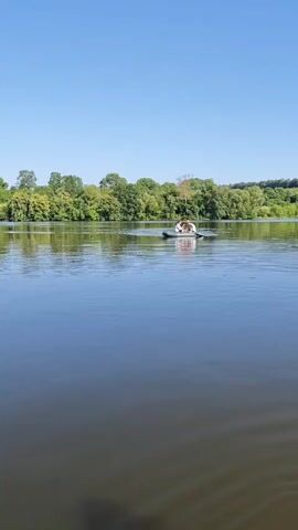 This media file shows a scenic view of a lake or wide river on a clear, sunny day. The sky is a vibrant, cloudless blue, indicating fair weather and likely midday or early afternoon. A dense line of lush green trees forms the backdrop along the far bank, suggesting a natural, possibly rural or park-like setting. The water is calm, with subtle ripples reflecting the blue sky and the green of the trees, creating a peaceful atmosphere.

In the middle of the water, a small, dark-colored inflatable boat is visible. Two people are in the boat, appearing to be enjoying a leisurely outing. One person, possibly steering or paddling, is wearing a white shirt and light-colored shorts or pants. The other person, in the front of the boat, is also wearing light-colored clothing and a hat. Their posture suggests they are relaxed and engaged in conversation or simply taking in the surroundings.

The overall mood is serene and idyllic, evoking a sense of calm and enjoyment of nature. The vibrant greenery and clear blue sky point to a warm season, likely summer. The presence of the boat and people suggests a recreational activity on the water. No visible text is present in the media file. The scene appears peaceful and idyllic, with the natural landscape dominating the view.