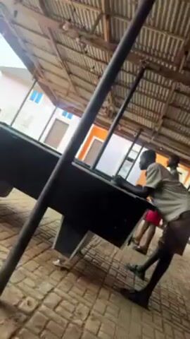 The video shows a man playing pool outdoors, under a corrugated metal roof supported by metal posts. The pool table is black with what appears to be a green playing surface. The man is wearing a light-colored shirt and dark shorts. The ground is paved with bricks. In the background, there are buildings with orange and white walls and blue windows. The camera angle is low, looking up at the scene. The camera moves slightly during the video.