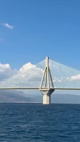 This is a video of the Rio–Antirrio Bridge, a cable-stayed bridge spanning the Gulf of Corinth, connecting the Peloponnese peninsula to mainland Greece. The video shows the bridge from a low angle, with the deep blue water in the foreground and the bridge stretching across the frame. The sky is a vibrant blue, dotted with fluffy white clouds, indicating a pleasant, sunny day. Mountains form a backdrop to the bridge, with a faint outline of a town or city visible along the coastline behind the water. The bridge itself is a striking architectural feat, with its tall, concrete pylons and the intricate web of white cables that support the roadway. The camera pans slowly across the bridge, offering a panoramic view of its impressive scale and the surrounding landscape. There is no visible text in the video. The overall mood is serene and awe-inspiring, highlighting the grandeur of the bridge and its natural setting.