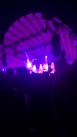 A low-angle shot looks up at a concert stage illuminated by purple lights. A crowd of people is visible in the foreground, silhouetted against the stage. The stage is set up with speakers and lighting equipment. A band is performing on stage, but their faces are not clearly visible due to the distance and lighting. The overall atmosphere is one of a lively music event.