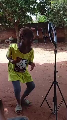 A young child wearing a green t-shirt with a Mickey Mouse graphic, gray leggings, and tan sandals is seen in a medium shot, dancing in front of a ring light mounted on a black tripod. The child is smiling and appears to be enjoying the dance. The background consists of a dirt ground, a brick wall, some laundry drying, trees, and other miscellaneous objects. The scene is set outdoors during the day. A glimpse of another person’s foot is visible in the lower left corner of the frame.
