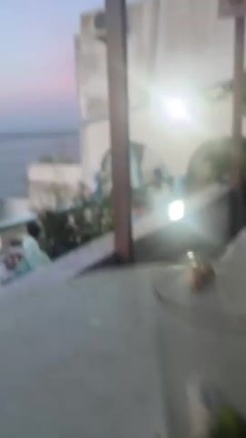 The video shows an outdoor terrace restaurant overlooking the sea. The sky is a gradient of pink, orange, and blue, indicating either sunrise or sunset. People are seated at tables, seemingly enjoying a meal or drinks. Some people wear head coverings, suggesting a culturally specific setting. The restaurant has a white and blue aesthetic, with some greenery visible around the perimeter. In the distance, another similar structure with people on its terrace can be seen. The camera pans slightly, showing different seating areas and views. Overall, the atmosphere appears relaxed and scenic.
