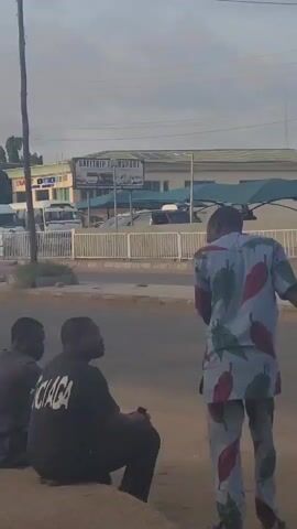 The video shows four men near a road in front of a building. Three of the men are sitting on a ledge. One of the men sitting is wearing a black shirt with the word "Balaga" on it. The fourth man is standing, facing the seated men. He is wearing a patterned outfit of white and blue with red leaf-like designs. In the background there is a street with vehicles and a building with a sign saying "SAFETRIP TRANSPORT". The sky is overcast.