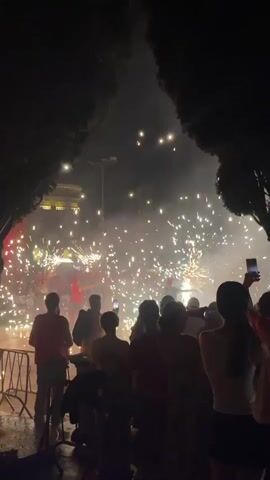 The video opens with a wide shot of a crowd of people gathered at night. Sparklers are being lit, creating a dazzling display of light and sparks. The crowd is illuminated by the fireworks and the lights from a stage in the background. The camera then pans to a woman's face, her eyes wide with wonder as she watches the spectacle. The video ends with a blurry shot of the crowd and the fireworks, capturing the energy and excitement of the event.
