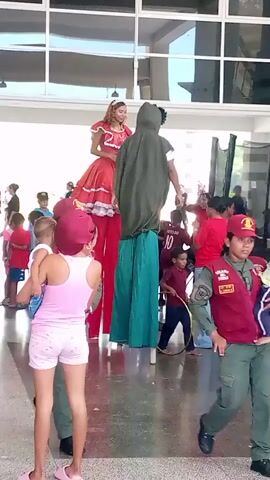 A group of people are gathered indoors in a bright, open space with large windows. In the center of the frame, two individuals are on stilts. The person on the left is dressed in a red, voluminous gown and appears to be a woman or girl. The person on the right is wearing a green hooded cloak over blue jeans and a white shirt. They are both looking downwards, interacting with the crowd. To the left of the stilt walkers, a young girl in a pink tank top and shorts is holding a baby. Another woman, wearing a red and gold uniform, possibly a security guard or event staff, is standing and looking towards the stilt walkers. To the right of the stilt walkers, a young girl in a red top and grey pants is running and smiling. Other people, including children and adults, are visible in the background, some watching the stilt walkers and others engaged in their own activities. The floor is reflective, suggesting a polished surface. The overall atmosphere appears festive and engaging.