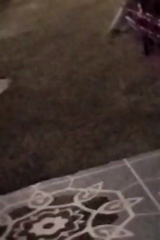 This is a video of a white cat sitting on a floor, looking towards a door. There is another white cat lying down in the background. The lighting is dim.