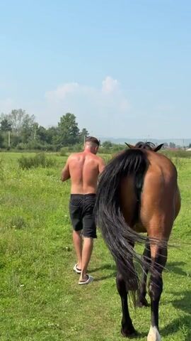 A shirtless man with short brown hair walks on a dirt path through a grassy field on a sunny day, holding a red rope attached to a brown horse walking beside him. The man is wearing black shorts and white flip-flops. The horse is facing away from the camera, and its tail is flicking. In the background, there are trees and some distant buildings.