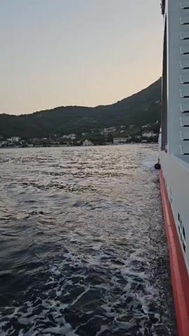 The video shows a view from a boat moving through water. The boat's side, which is white with a red stripe, is visible on the right side of the frame. The water is choppy, creating white foam and wakes as the boat moves forward. In the background, a hilly coastline with houses and trees is visible. The sky is a pale gradient of blue and gray, suggesting it is either early morning or late evening. The overall impression is of a serene journey across the water, with a scenic landscape in the distance.