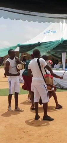 A group of people are gathered outdoors under a large tent. In the foreground, two young men are playing drums. A young woman in a green shirt and red skirt is dancing. Behind them, a group of women dressed in similar attire are also dancing, holding white feathers. Spectators are visible in the background, seated under the tent. The ground is sandy. The sky is overcast.