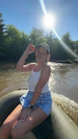 A young woman with brown hair, wearing a white tank top, light blue shorts, and a green baseball cap, sits in a dark green inflatable boat on a river. The sun shines brightly behind her, creating a lens flare. She initially shields her eyes from the sun with her right hand, then smiles at the camera, then looks to her right. Trees line the riverbank in the background. The boat is moving, creating a wake behind it.
