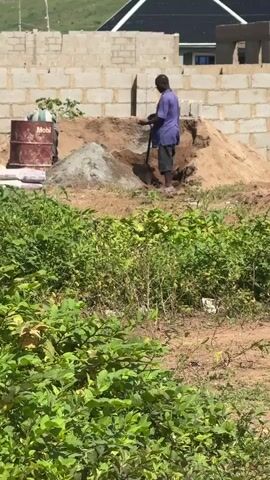 A man in a purple shirt and shorts is using a shovel to dig into a pile of sand. Next to him is a red barrel and several bags of cement. Another person is partially visible on the left side of the frame, also near the barrel. The background shows a partially constructed building with cinder block walls and a dark roof. Green foliage is in the foreground and surrounding the construction site.
