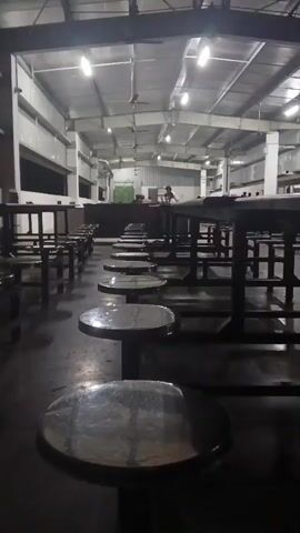 The video shows a large, open cafeteria or dining hall. Rows of tables and stools are visible, most of them empty. The lighting is artificial, coming from overhead fixtures. In the background, several people are seated at tables, some appearing to be eating or talking. The overall impression is that of a functional, possibly industrial, dining area during a period of low activity.