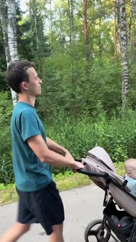 A young man with short brown hair and wearing a teal t-shirt and black shorts walks on a paved path through a wooded area. He is pushing a beige stroller with a baby inside. The baby is wearing a light blue shirt and has blonde hair. The path is surrounded by lush green foliage and trees. The lighting suggests it is daytime, possibly late afternoon.