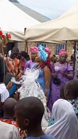 Here is a concise description of the video:

The video shows a group of people gathered under a tent, seemingly at an event. The focal point is a woman in a blue dress and a light blue headwrap, standing near a large, wrapped gift. Several people are dressed in matching purple outfits with pink headwraps. A man is playing a drum. The crowd consists of both adults and children.  The environment appears to be outdoors, with a building visible in the background.