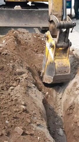 An excavator is digging a trench. The excavator's bucket is in the trench, scooping up dirt and then lifting it out and depositing it on the side of the trench. The trench is narrow and deep, with loose soil piled up on either side. The excavator is yellow and appears to be in good working condition. There are no people visible in the immediate vicinity of the excavator, but a person can be seen standing in the background, near a sign. The environment appears to be a construction site, with a road or paved area visible in the background. The lighting suggests it is daytime.
