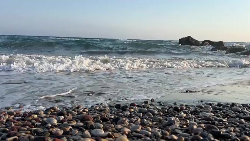 This is a video of a gentle wave rolling onto a pebble beach. The foreground is filled with smooth, rounded pebbles of various colors, including grey, brown, and white. In the middle ground, the ocean is calm, with small waves breaking and creating white foam as they reach the shore. The water is a deep blue-green. In the background, larger rocks are visible in the sea, and the horizon is a pale blue sky.

The time of day appears to be late afternoon or early evening, judging by the soft, warm light. The weather seems pleasant, with a clear sky and calm seas. The setting is clearly a beach, and the presence of pebbles suggests a natural, possibly undeveloped coastline. The overall mood conveyed by the video is peaceful and serene, evoking a sense of tranquility and the gentle rhythm of nature. There is no visible text in the video.