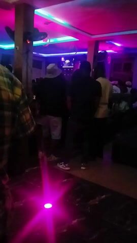 The interior of a dimly lit bar or club is visible in the video. Several people are milling about. The lighting is provided by pink, blue, and green lights in the ceiling, as well as a spotlight that cycles through pink, orange, and blue. There are ceiling fans, and a TV screen is visible in the background. The floor is patterned, and there are chairs and tables in the distance.