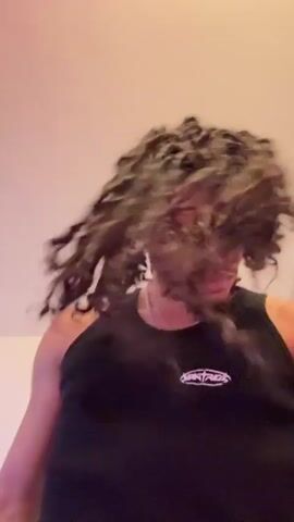 A man with dreadlocks is facing the camera. He is wearing a black tank top with a small, light-colored logo on the left breast. He has a silver chain around his neck and a tattoo on his neck. He appears to be inside a room with white walls. He shakes his head, causing his hair to move rapidly. There's a white earbud in his right ear. Clothes are piled on a bed behind him.