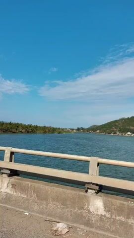 The video shows a first-person perspective of riding a motorbike on a bridge. The bridge has grey concrete railings, and there are tall, red flags attached to the railing at intervals along the length of the bridge. The camera moves along the bridge, showing the view of the water and shoreline on either side. The sky is blue with scattered clouds, and there are some trees and buildings visible along the shoreline. A few trucks are visible driving in the opposite direction.