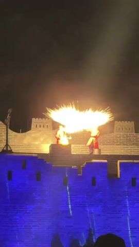 At night, a performance stage with the set looking like part of a wall is illuminated in a blue hue. Two figures dressed in red Chinese-style costumes are seen on the wall, one standing near a small fire pit. They stand on the structure while a large display of gold-colored fireworks explodes above them. The backdrop is a dark night sky.