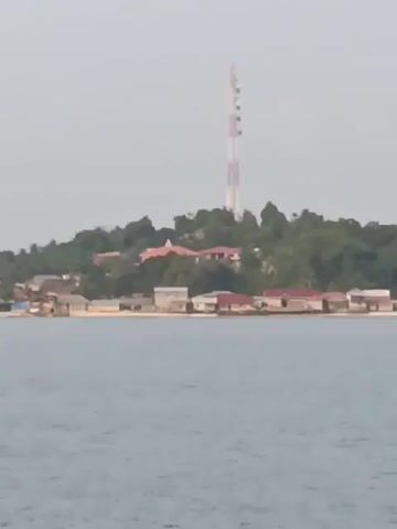 The video shows a body of water, likely the sea, with a coastline in the background. On the coastline, there are several small buildings and a tall radio tower on a hill. The sky is overcast. The video appears to be taken from a moving boat. There are no people or animals visible in the frame.