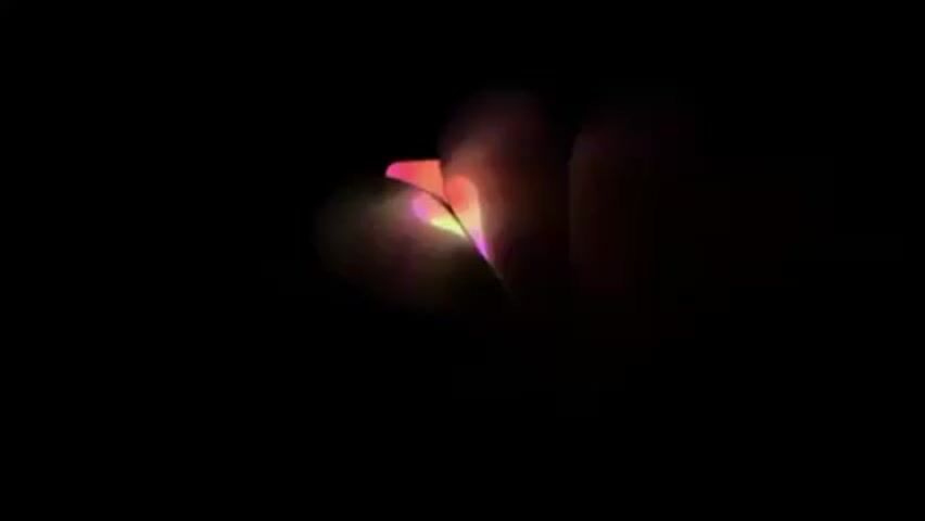 In a very dark scene, a hand is making a heart shape by bending the fingers.  A pink and yellow light is shining through the fingers, highlighting the heart shape.