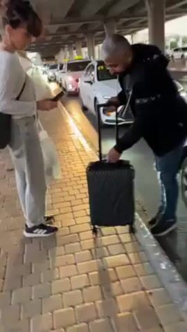 A man standing on a bricked sidewalk, opens the trunk of a white car and puts a black suitcase in the trunk. The man is wearing jeans and a black jacket with white details and black shoes. The car is parked underneath an overpass and there are other cars in the background. Another person is standing beside him, obscured.