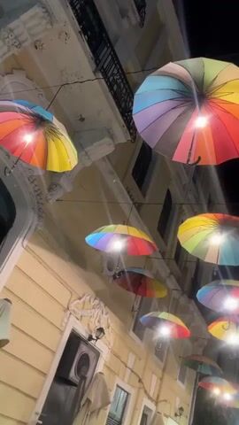 The video captures a night scene on a narrow street in Karditsa, Greece. Numerous rainbow-colored umbrellas are suspended overhead, forming a canopy across the street. Each umbrella appears to have a light source attached beneath it, illuminating the umbrellas themselves and casting a warm glow downwards. The sky above is dark, indicating nighttime. Tall, light-colored buildings with ornate details, balconies, and windows line both sides of the street, suggesting an urban or town setting. The lights from the umbrellas and some street-level fixtures illuminate the facades of the buildings. No people or vehicles are visible, and there is no discernible activity. The overall atmosphere is whimsical and decorative.