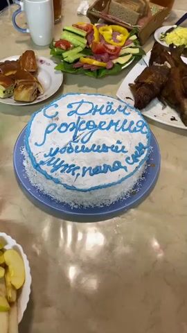 The scene is a close-up shot of a table laden with food, presumably for a birthday celebration. The focus is on a round cake decorated with white frosting and blue lettering, which reads "С Днём Рождения любимый муж, папа!", translating to "Happy Birthday, beloved husband, dad!". The cake is on a light blue plate. Surrounding the cake are plates of food, including what appears to be smoked fish, a platter with sliced cucumbers, tomatoes, yellow bell peppers, and sausage, rolls filled with various ingredients, and a bowl with sliced apples. A mug is also visible in the upper left corner. The table has a glossy, light brown surface. There are no visible people in the frame.