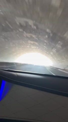 The video shows a point-of-view shot from inside a car driving through a tunnel and then onto a highway. The car is moving forward. In the first few seconds, the car is in a dimly lit tunnel, with the exit visible as a bright light in the distance. The tunnel walls appear to be made of stone or concrete. As the car exits the tunnel, the scene changes to a sunny day with a blue sky and white clouds. The car is on a paved highway with multiple lanes. There are overhead signs with some text on them, and to the right of the highway, there is a yellow sign with text and graphics. Trees and hills are visible in the background. The interior of the car is visible in the foreground, showing a leather dashboard with a diamond-stitched pattern and a blue illuminated section. The video is short, capturing a transition from a dark tunnel to an open road.