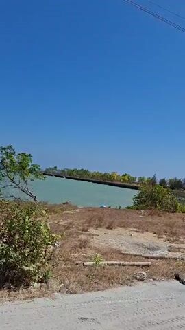 The video opens with a clear blue sky and a body of water. The camera pans across a flatbed truck loaded with metal plates and chains, then to a black truck with graphics on its side, followed by a rusty truck with a wooden bed. The camera then shows a large green tree with a yellow barrier in front of it, and finally, a shot of the blue sky. The footage appears to be taken from a moving vehicle.