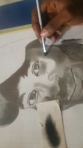 The video shows a close-up of a hand holding a fine-tipped paintbrush and applying shading to a charcoal drawing of a Black man's face. The drawing is done on white paper with a grid faintly visible underneath. A piece of paper is used as a shield to protect areas of the drawing from smudging. The hand is carefully shading the area around the left eye of the portrait, adding detail and depth. The drawing is laid out on a blue surface. The video is shot from a slightly elevated perspective.