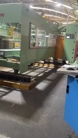 The video shows a large, light green industrial machine in a factory setting. The camera moves slowly along the side of the machine, focusing on its lower section, which is equipped with a series of windows. Through the windows, internal mechanisms of the machine are visible, including what appears to be a stack of materials. The floor around the machine is dark and industrial, with some yellow safety markings. To the right of the machine, there's a blue cabinet and a desk with a computer mouse and papers. The overall impression is of a controlled industrial environment.