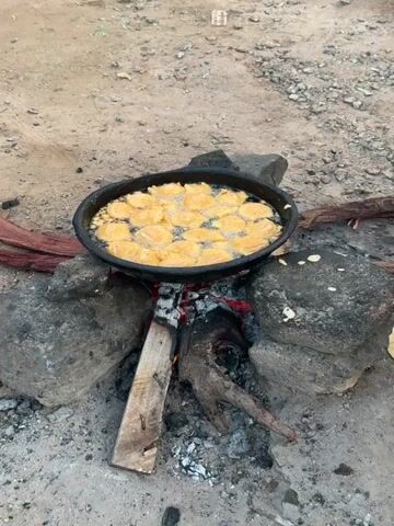 The media file shows a close-up view of a cooking scene set outdoors. A dark, round frying pan, filled with numerous golden-brown, fritter-like food items, is placed over a small, smoldering fire. The fire is built using logs and rocks, with embers glowing red beneath the pan. The surrounding ground is rough and appears to be made of dirt and gravel, typical of an outdoor or rural setting.

To the right of the pan, there are several large, dark rocks supporting the cooking setup. A flat, circular, light-colored object, possibly a type of dough or uncooked food item, rests on the ground next to one of the rocks. Further to the right, a blue plastic bowl and a partially visible red and gray bucket containing food are also present, suggesting that ingredients or prepared food are being kept nearby. A person's patterned fabric-covered leg is visible in the bottom right corner of the frame, indicating human presence and activity.

The lighting suggests it could be daytime, possibly late afternoon or early morning, given the ambient light. There are no explicit indicators of weather, but the scene is exposed to the elements. The overall impression is one of traditional or rustic outdoor cooking, likely in a village or rural area. The focus is on the frying process, with the food items bubbling in the hot oil. No text is visible in the media file.