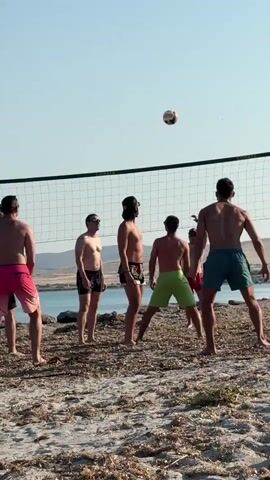 In this horizontal video, a group of men are playing beach volleyball. The game is being played on a sandy beach, with the sea and hills visible in the background. The sky is clear and blue, suggesting a sunny day. The men are all wearing swim trunks and appear to be enjoying themselves. They are spread out across the court, with one man in the foreground on the left, and another in the foreground on the right, facing the net. The ball is in the air, just above the net, indicating that the game is in progress. The overall mood is casual and fun, typical of a day at the beach. There is no visible text in the video.
