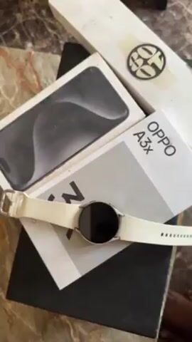 The video shows an arrangement of several boxes and a smartwatch on a dark marble-patterned surface. The boxes are white and black. The boxes appear to be packaging for electronics: a white box labelled "OPPO A3x," a white box with a blurry image of what could be an iPhone, and a longer white box labelled "Samsung Galaxy Watch6". A smartwatch with a black screen and a white band sits on top of the boxes. The background is blurry and indistinct.