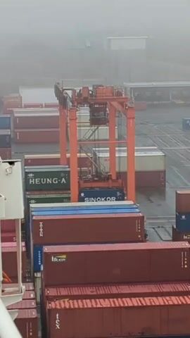 The video shows a shipping container yard on a foggy day. A large orange gantry crane is positioned over several stacks of shipping containers. The crane appears to be in operation, with its lifting mechanism suspended above a blue container labeled "SINOKOR". Other containers are visible in the background, some with labels such as "HEUNG-A" and "TEX". The ground is wet, suggesting recent rain or high humidity. The overall environment appears industrial and operational.