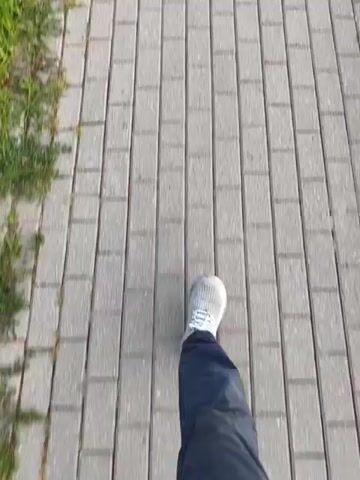 A first-person perspective video shows a person walking along a paved sidewalk. The sidewalk is made of gray rectangular bricks laid in a pattern. To the left of the sidewalk is a strip of green grass and some small bushes. To the right of the sidewalk is also green grass and a road beyond that. In the distance, trees and utility poles can be seen. The sky is a pale orange and yellow, suggesting either sunrise or sunset. A tall electrical tower is visible in the far distance.