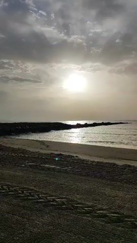 The video shows a beach scene with the sun setting over the ocean. There is a breakwater made of rocks extending into the water, and waves are gently lapping at the shore. The sand in the foreground has tire tracks, indicating that a vehicle has been on the beach. The sky is filled with clouds, but the sun is breaking through, casting a warm glow on the water. The overall mood is peaceful and serene.