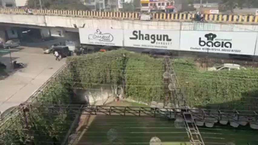 The video shows a view from an elevated position overlooking a street scene in what appears to be a town or small city. There is an overpass with large signs for "Shagun," "Bacia," and "Amar Palace." Below the overpass, a road with some vehicular traffic is visible. There are buildings on either side of the street. In the foreground, there is a green, possibly landscaped area enclosed by fencing. The sky is overcast and the general environment suggests a daytime setting.